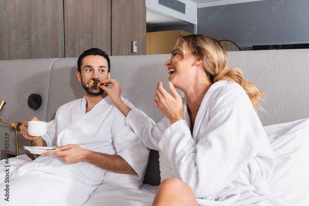 Cheerful woman feeding croissant to muslim boyfriend with coffee on hotel bed Cheerful woman feeding croissant to muslim boyfriend with coffee on hotel bed