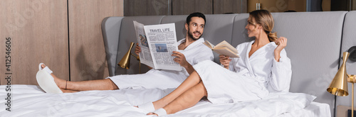 Smiling interracial couple with newspaper and book sitting on hotel bed, banner