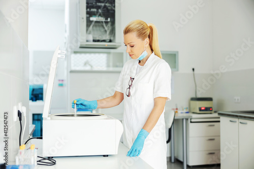 Middle aged experienced lab assistant with rubber gloves and face mask on putting test tube with vaccine in machine. Research for cure for corona virus.