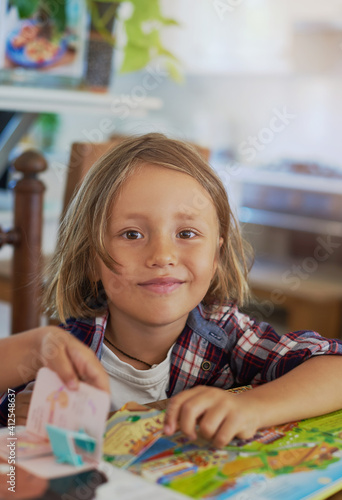 Portrait of joyful little boy which poses sitting at table with books and looks at camera in living room.