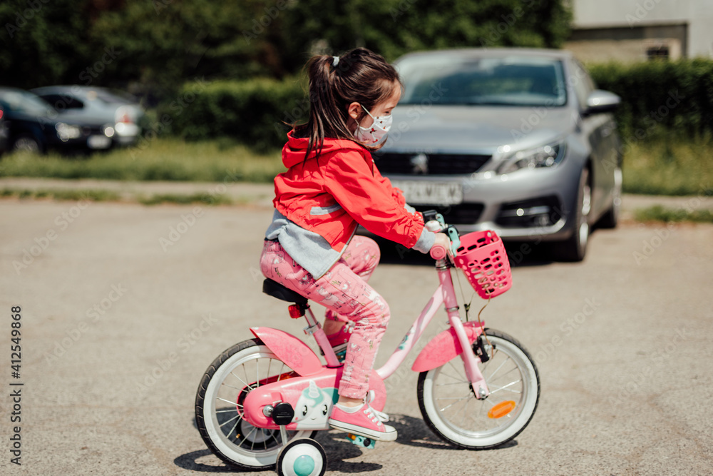 a little girl in red with a face mask outdoor walking on a bicycle after coronavirus covid 19 started