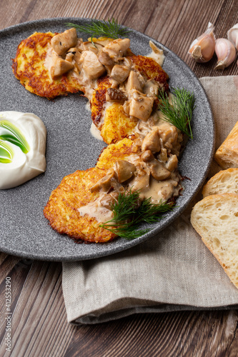 Fried potato pancakes served with mushrooms, sour cream and dill on a grey round plate. Close-up isolated on wooden background with grey linen napkin, sliced bread and garlic.