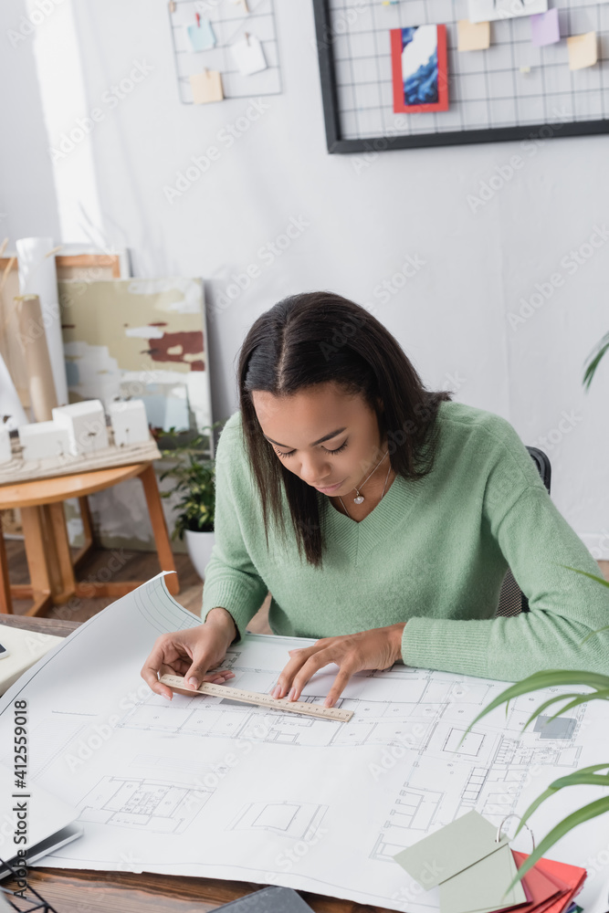 concentrated african american architect measuring blueprint with ruler at workplace
