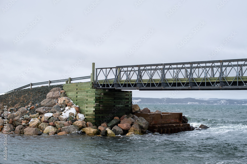 A large metal footbridge spans over a stream.  The background is a cloudy sky with some blue breaking through.  The bailey bridge is metal.  The ocean is in the background with rough seas. 