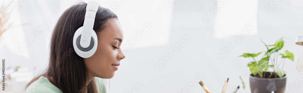 Naklejka premium side view of smiling african american interior designer listening music in wireless headphones, banner