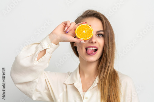 Portrait of a cheerful caucasian young woman covering eye with an orange slice and stick out tongue wears white shirt against a white background