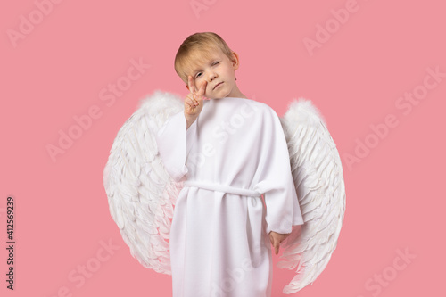 Angel with wings in white clothes stand showing selfie or peace sign looking into camera. Studio shot on pink background.