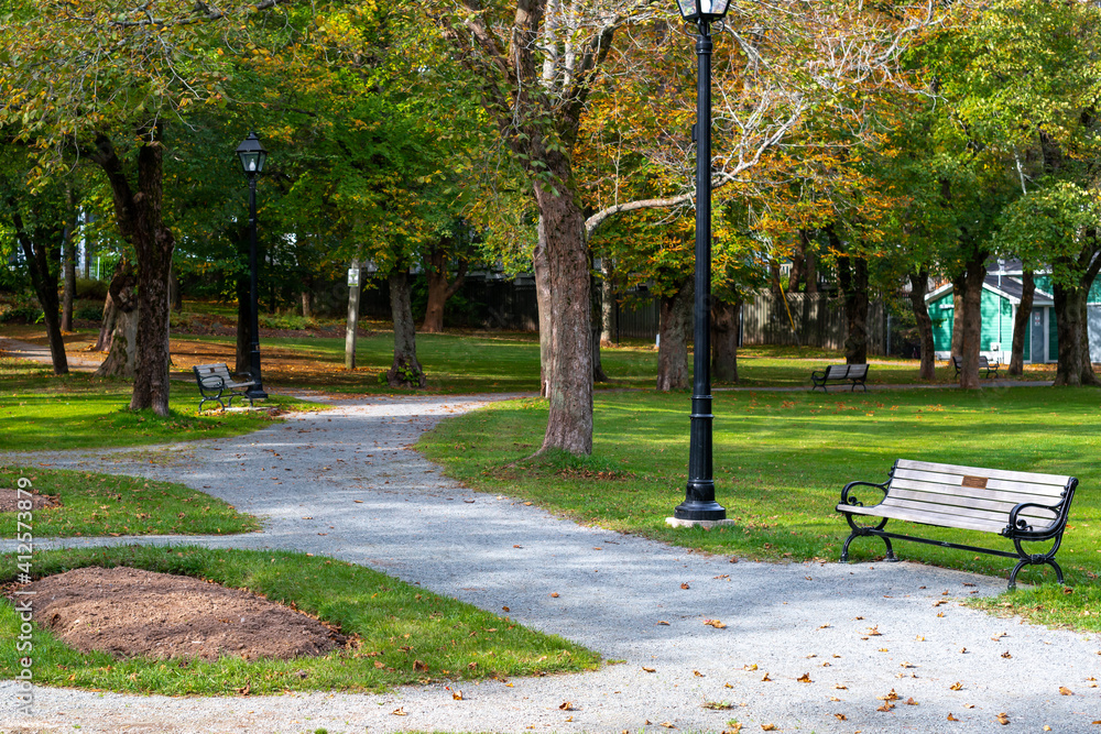 Obraz premium A long gravel footpath in a park with a bench and lots of tall trees. At the end of the trail is a red brick building. The sky is blue and the grass is very green. There are leaves on the tall tree