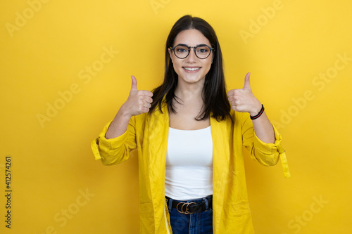 Young brunette businesswoman wearing yellow blazer over yellow background success sign doing positive gesture with hand, thumbs up smiling and happy. cheerful expression and winner gesture.