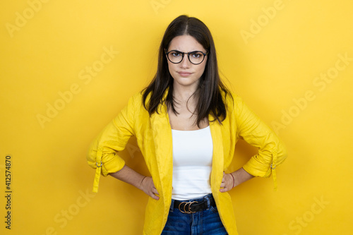 Young brunette businesswoman wearing yellow blazer over yellow background skeptic and nervous, disapproving expression on face with arms in waist