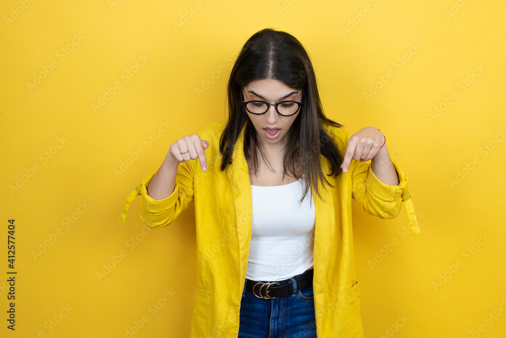 Young brunette businesswoman wearing yellow blazer over yellow background surprised, looking down and pointing down with fingers and raised arms Young brunette businesswoman wearing yellow blazer over yellow background surprised, looking down and pointing down with fingers and raised arms