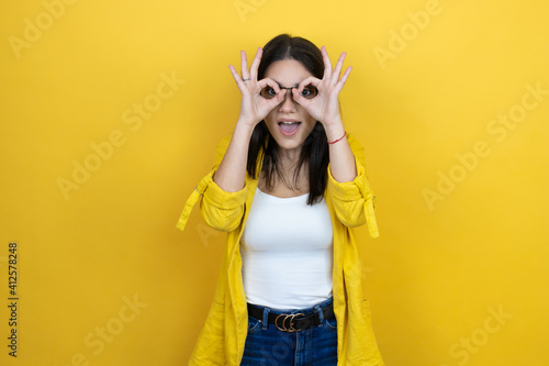 Young brunette businesswoman wearing yellow blazer over yellow background doing ok gesture shocked with smiling face, eye looking through fingers