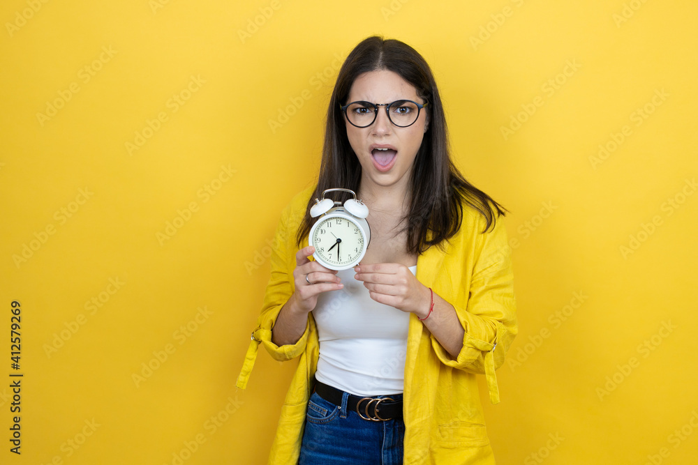 Fototapeta premium Young brunette businesswoman wearing yellow blazer over yellow background screaming and scared, pointing the clock