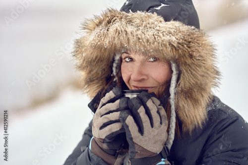 Happy smiling woman wearing a fur trimmed hooded jacket enjoying a mug of coffee cradled in her gloved hands outdoors in winter snow in a concept of the seasons in a close up portrait