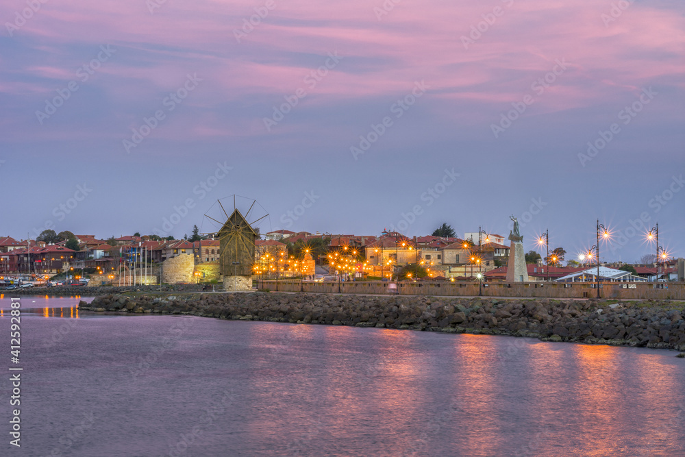 Old windmill and citylights at colorful sunset.  Amazing view of the windmill, located on a narrow man-made isthmus at the entrance of the UNESCO - protected ancient town of Nessebar, seaside resort.