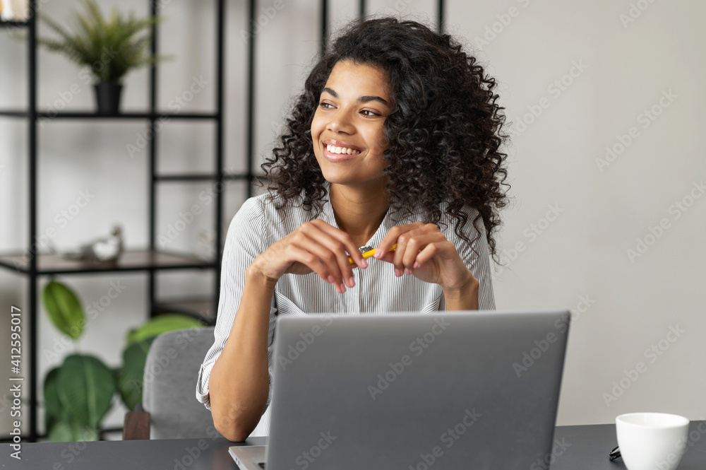 Pretty charming young African American woman with Afro hairstyle sitting at the desk and looking in the window, taking a break from working on a laptop, dreaming about vacation, enjoying her workplace Pretty charming young African American woman with Afro hairstyle sitting at the desk and looking in the window, taking a break from working on a laptop, dreaming about vacation, enjoying her workplace