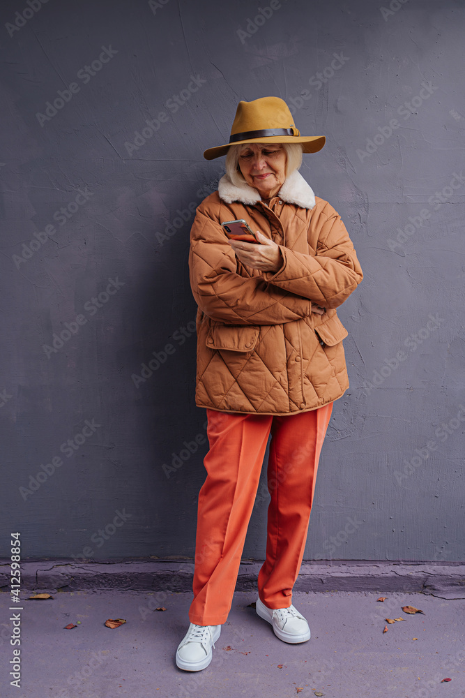 Stylish senior woman in warm clotheswith smartphone. Full body of confident aged female in trendy outerwear and hat while standing near gray wall. Stylish senior woman in warm clotheswith smartphone. Full body of confident aged female in trendy outerwear and hat while standing near gray wall.