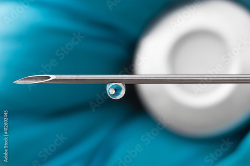 Tip of a cannula with a drop of vaccine and bottle with septum in a hand with blue vinyl protective glove macro