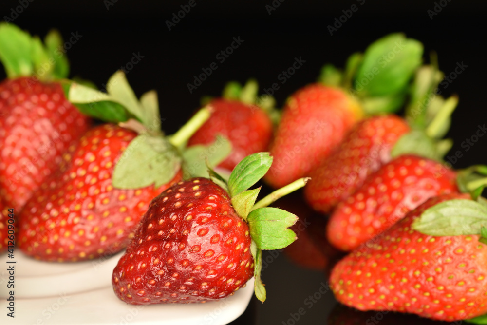 Several berries of bright red ripe organic tasty strawberries on a white ceramic saucer. The background is black. Several berries of bright red ripe organic tasty strawberries on a white ceramic saucer. The background is black.