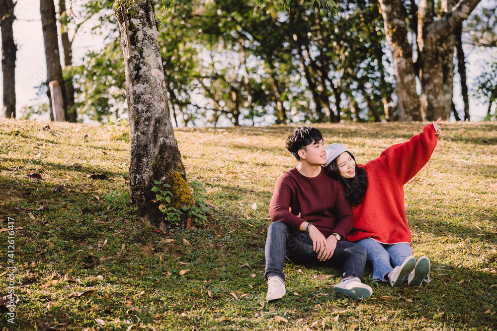 Portrait sweet couple of teenage sitting on hill, asian man with maroon sweater hug or cuddle a young woman with red sweater. Romantic mood and sweet emotional relationship in summer with copy space Portrait sweet couple of teenage sitting on hill, asian man with maroon sweater hug or cuddle a young woman with red sweater. Romantic mood and sweet emotional relationship in summer with copy space