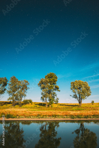 Blue Night Starry Sky Above Lake River. Night Glowing Stars And Trees Woods Reflected In Water. Night Landscape