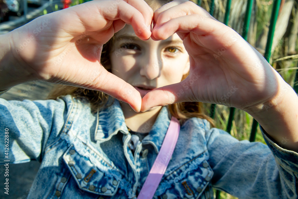 Fototapeta premium teenage girl with long hair looks through folded fingers in the shape of a heart on a sunny day