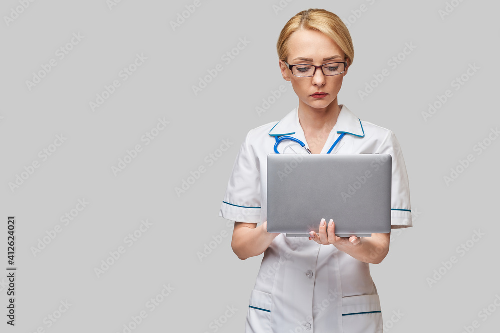 Beautiful Caucasian woman doctor or nurse holding a laptop pc computer standing over grey background Beautiful Caucasian woman doctor or nurse holding a laptop pc computer standing over grey background