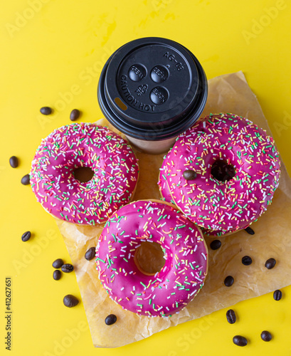Yummy pink glaze donuts with colorful sprinkles, takeaway coffee cup and roasted coffee beans on yellow background. Junk food. Top view.