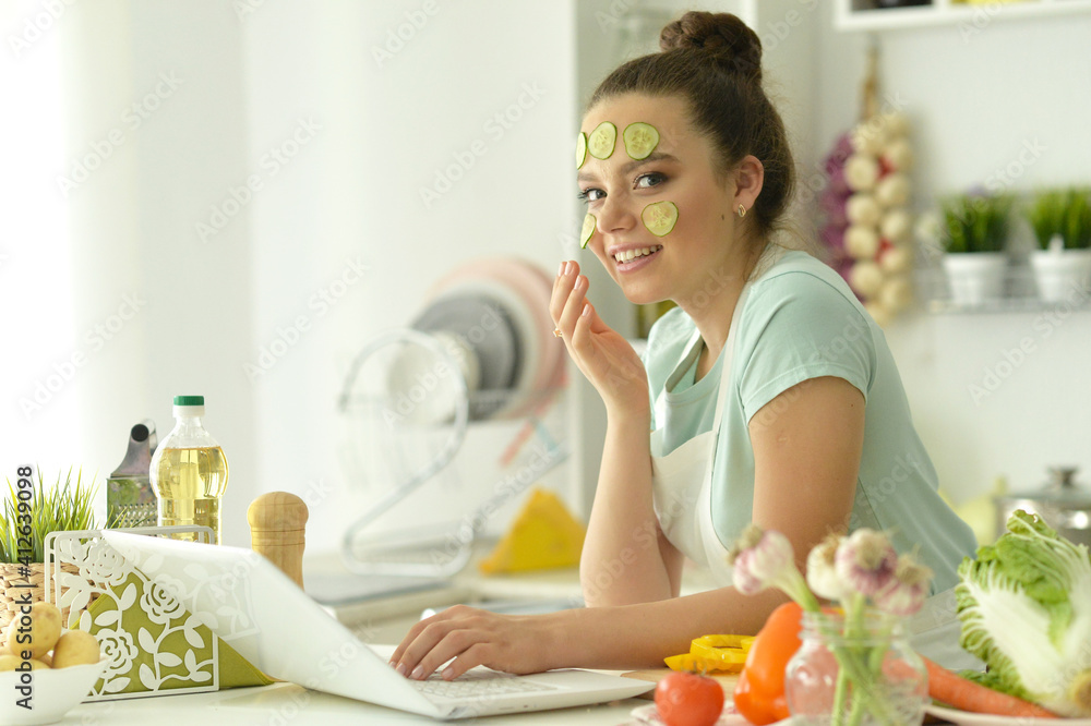 Beautiful young woman making salad and using laptop at home