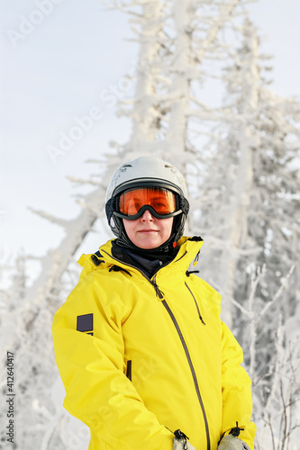 Cute young woman snowboarder in a bright overalls in a sports helmet and ski goggles on the background of a winter forest. Extreme sports. Vertical shot