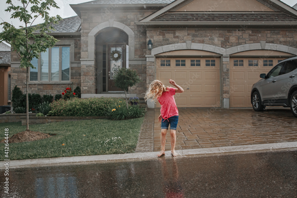 Cute adorable girl splashing under rain in front of house. Child having fun during rain shower storm. Seasonal summer outdoors activity for kids. Freedom and happy childhood lifestyle.
