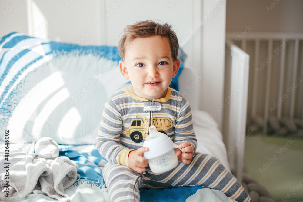 Cute adorable Caucasian kid boy sitting on bed drinking milk from kids