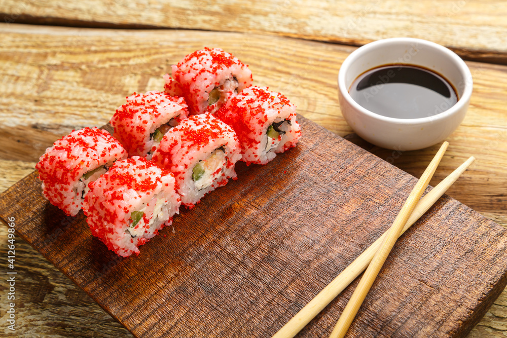 Roll with shrimp cucumber and red tobiko caviar and sticks on a board and soy sauce in a sauce bowl on a wooden background. Roll with shrimp cucumber and red tobiko caviar and sticks on a board and soy sauce in a sauce bowl on a wooden background.