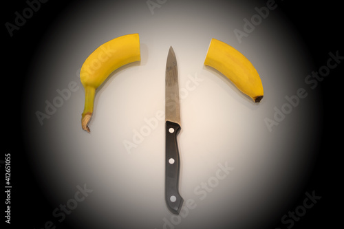 Banana is sliced in half on a dark background. The knife lies in the middle between the two halves of the banana