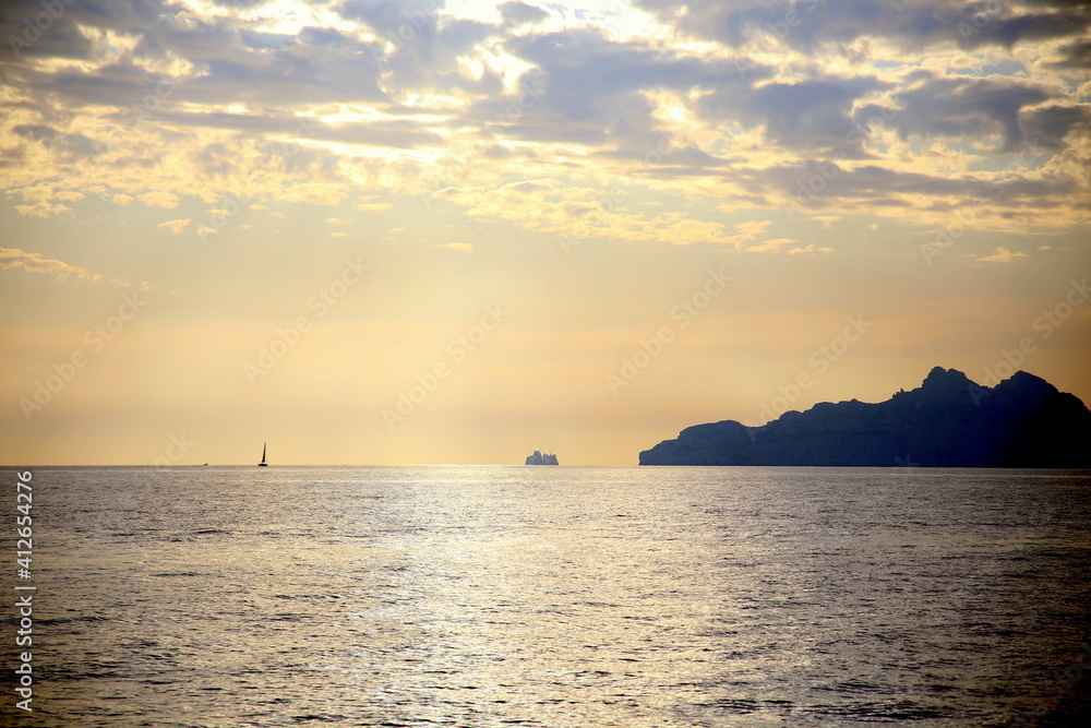 Sunset light on an isolated sailboat, sailing on the horizon, with the coast in backlight, under a dramatic sky