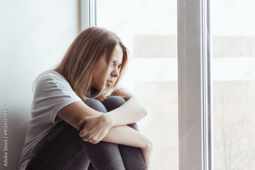 A young blonde beautiful despaired woman introvert in the white t-shirt and grey leggings is sitting on the windowsill, hugging her legs and looking out the window. Self-isolation, lock down. Solitude A young blonde beautiful despaired woman introvert in the white t-shirt and grey leggings is sitting on the windowsill, hugging her legs and looking out the window. Self-isolation, lock down. Solitude