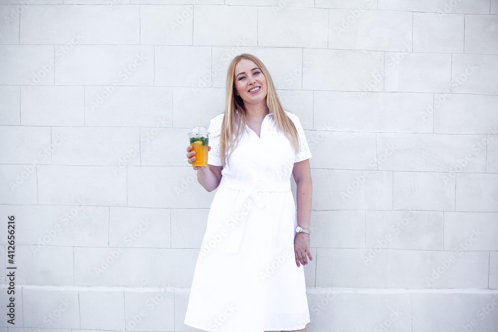 Beautiful overweight lady in white dress and holding the bagpack waking in the street of the city and smiling. Plus size girl drink orange cocktail against the white wall Beautiful overweight lady in white dress and holding the bagpack waking in the street of the city and smiling. Plus size girl drink orange cocktail against the white wall