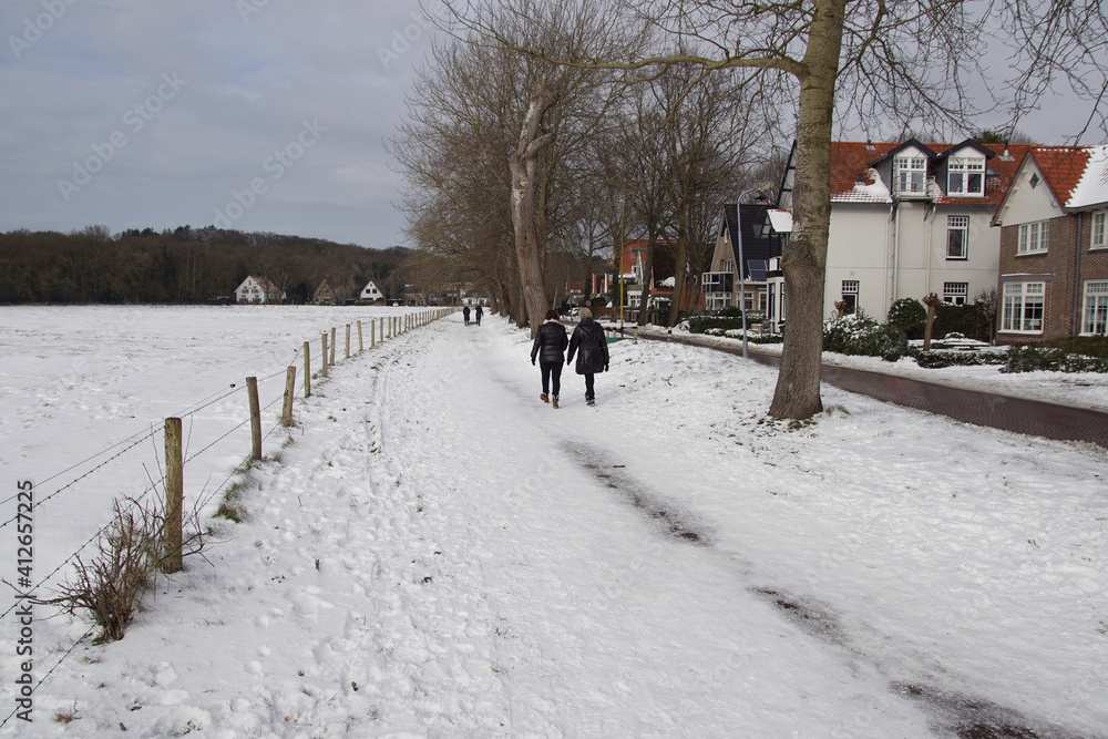 Street, footpath, trees, and meadow covered with snow in winter. Hikers in the Dutch village of Bergen. Forest in the distance. Netherlands, February. Street, footpath, trees, and meadow covered with snow in winter. Hikers in the Dutch village of Bergen. Forest in the distance. Netherlands, February.