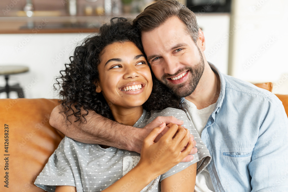 Hilarious multiracial couple in love sits in embraces in cozy living room at home. A close-up portrait of smiling caucasian guy and charming african girl are hugging. Love and affection concept Hilarious multiracial couple in love sits in embraces in cozy living room at home. A close-up portrait of smiling caucasian guy and charming african girl are hugging. Love and affection concept