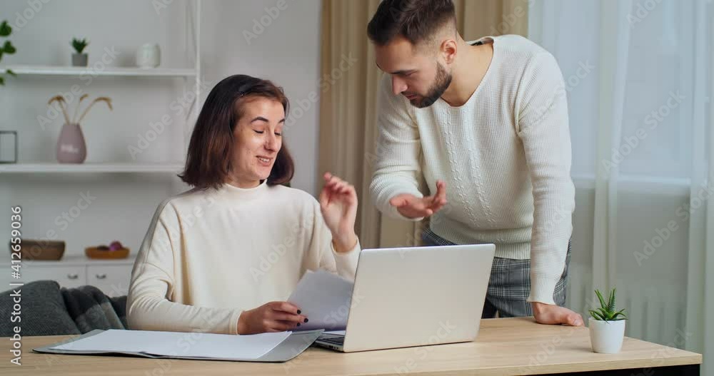 Young woman wife sits at home table looks at laptop has problems with paperwork makes mistake in document serious man husband approaches argues indignant reads letter feels anger on silly girl Young woman wife sits at home table looks at laptop has problems with paperwork makes mistake in document serious man husband approaches argues indignant reads letter feels anger on silly girl