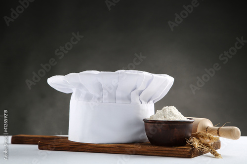 Chef hat with bowl of flour, wooden rolling pin and cutting board on black background