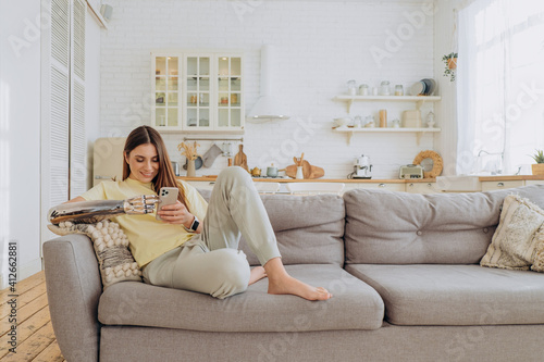Attractive woman with a bionic arm sits at home on the couch in comfortable home clothes with a phone and surfs the Internet or chat with friends online on a sunny day against the background of a