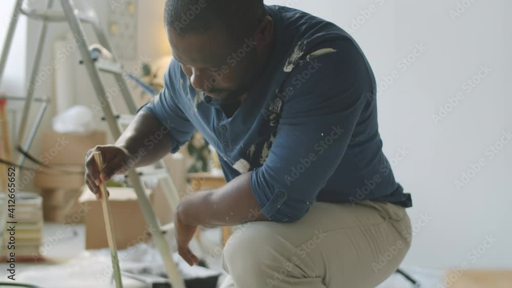 African American man stirring green interior paint in bucket and then pouring it into tray while renovating room in his apartment African American man stirring green interior paint in bucket and then pouring it into tray while renovating room in his apartment