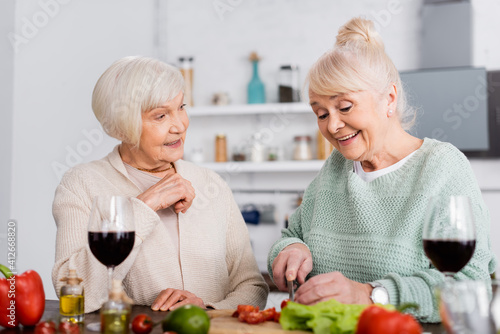 happy senior woman looking at joyful friend cutting vegetables in kitchen