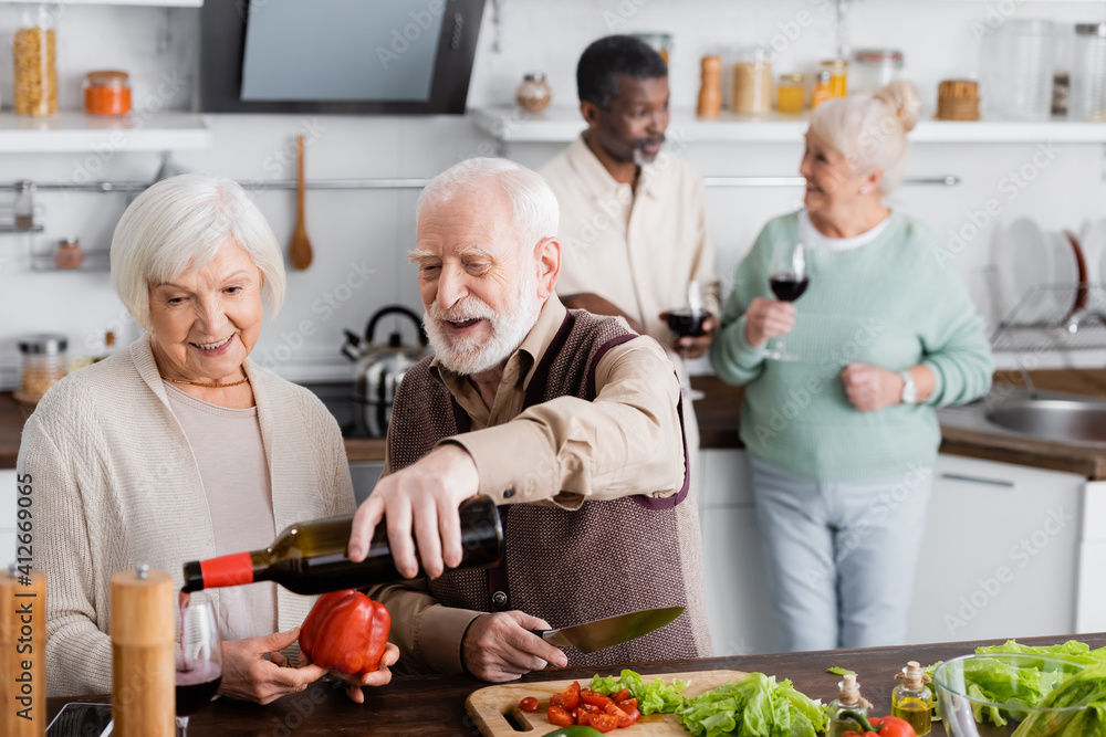 happy senior man pouring wine in glass near retired woman and multicultural friends on blurred background happy senior man pouring wine in glass near retired woman and multicultural friends on blurred background
