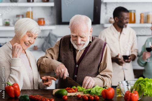 happy senior woman pointing with finger at retired man cooking salad near multicultural friends on blurred background