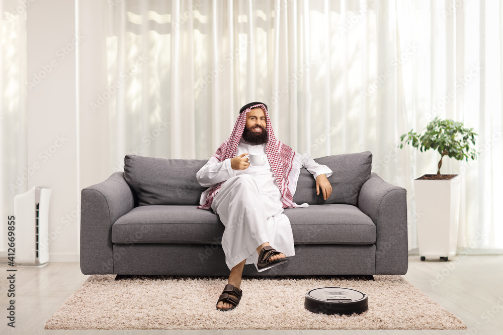 Arab man at home resting on s sofa and a robotic vacuum cleaner cleaning the floor Arab man at home resting on s sofa and a robotic vacuum cleaner cleaning the floor