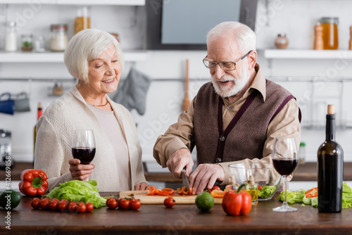 happy senior man cutting vegetables near smiling wife with glass of wine