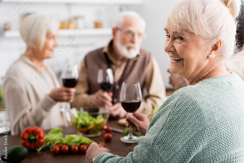 happy senior woman holding glass of red wine near retired friends on blurred background