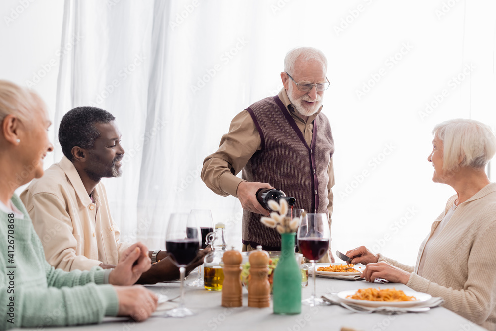 senior man in eyeglasses pouring red wine in glass near happy multicultural friends senior man in eyeglasses pouring red wine in glass near happy multicultural friends